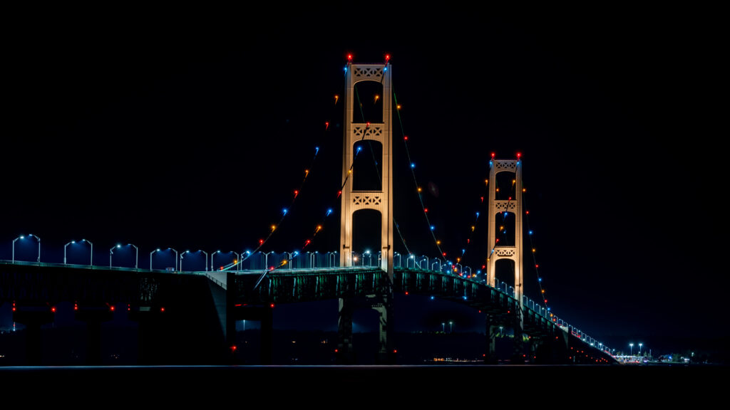 Night view of the Mackinac Bridge illuminated by colorful lights, photographed with long exposure to highlight detail and clarity in low light.
