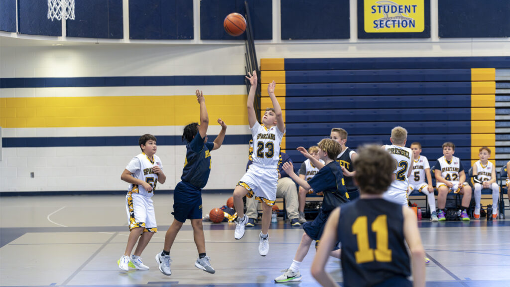 Wide indoor basketball action with multiple players jumping for a shot, photographed at a fast shutter speed using Auto ISO to freeze motion under uneven gym lighting.